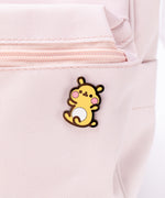 The Cheek pin in it’s packaging, laying on a white floor, a pair of pink and white peonies partially visible in the top left corner. The packaging is a square cardboard backing, a mint square in the middle and pink banners on the top and bottom. The top banner features the Pusheen Shop logo, and the bottom banner says ‘Cheek the Hamster Pin’ in white.