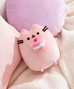 Cherry Plush lies on a white blanket in front of pink and purple pillows. Pusheen's facial features are shown in dark brown.
