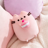 Cherry Plush lies on a white blanket in front of pink and purple pillows. Pusheen's facial features are shown in dark brown.