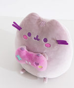 Gamer themed cat plush sits on a white desktop. The purple-hued cat holds a pink gaming controller with multi-colored buttons.