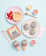 A hand holdind the corner of the Pusheen cook book, standing on top of a wooden surface surrounded by a rolling pin and a pink frosted cupcake.