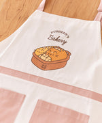Pusheen Apron lies on a wooden tabletop. Printed in the graphic on the chest of thw apron is Pusheen the Cat taking the form of a baguette, loaf of bread, and croissant sitting in a brown bread basket. 
