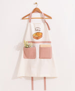 White and dusty pink apron hangs on a clothing hanger. A bundle of flowers and a notebook are placed in the oversized pink pockets on the front of the apron.