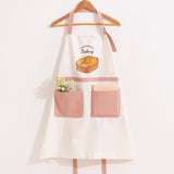 White and dusty pink apron hangs on a clothing hanger. A bundle of flowers and a notebook are placed in the oversized pink pockets on the front of the apron.