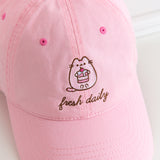 Close-up view of Pusheen hat lying on a white surface. The cap curved brim points towards the bottom right corner. In the center of the forehead section is an embroidery of Pusheen holding a cherry tiered cake.