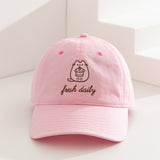 Pusheen Sweets Cap on a white surface. The light pink cap features Pusheen the Cat in a pink hue while outlined by a dark brown color. The phrase "fresh daily" sits below the cat embroidery. 