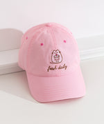 Top-down view of the Pusheen Sweets Cap. The top button and seams can be seen from this view as well as the thread detailing on the bill of the hat. The hat is propped on a white ledge to show the full size of the baseball-style cap. 