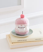 Pink plush toy resembling a macaron with a cherry on top, sitting on a stack of books.