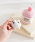 Model holds the removable Pusheen the Cat white plush that sits inside the cherry macaron plush environment. The light plush rests between the model’s fingers.  