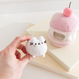 Model holds the removable Pusheen the Cat white plush that sits inside the cherry macaron plush environment. The light plush rests between the model’s fingers.  