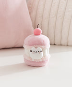 Cat plush toy resembling a macaron with a cherry on top, sitting on a light pink surface. A white Pusheen the Cat mini plush is able to removed from the macaron plush environment. 
