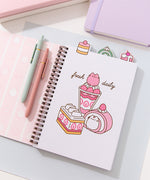 A light pink spiral bound notebook rests on  a white stair display. The large graphic on the front cover shows Pusheen as four different desserts including cakes, a macaron, and a swiss roll.