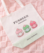 Pusheen Sweets Tote Bag lies on white chair. The white canvas tote bag features a grey Pusheen the Cat inside three sweet dessert macarons. Above the macaron graphic is the phrase “Pusheen Macarons”  and below the graphic is the phrase "sweet moments"