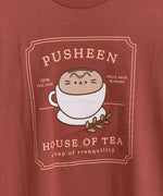 Red brown graphic tee lies on a white tabletop. The tee has a large graphic in the center of the tee featuring Pusheen the Cat in a tea cup.