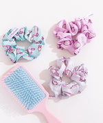 Pusheen 3-Pack Hair Scrunchies held together on their cardboard backing card loop. The top half of the card has the Pusheen logo in brown on a light pink background and the bottom half of the card is purple.  