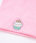 Light pink Pusheen beanie hat laid on a white surface accompanied by cupcakes with white, yellow, and pink frosting. 