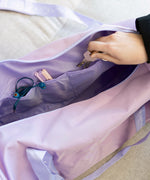 Pusheen Gym Bag sits on a turf surface with a water bottle. The purple bag has a velcro and zippered pocket on one side for easy access storage. 
