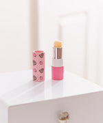 Unscented Pusheen lip balm sits on a desktop. The clear lip balm has pink packaging with strawberry graphics. 