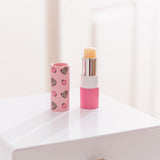 Unscented Pusheen lip balm sits on a desktop. The clear lip balm has pink packaging with strawberry graphics. 