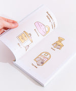 The back of the Pusheen the Cat book resting on top of a white desk. The back cover features copy, illustrations of Pusheen floating with a balloon and Pusheen in front of a computer. The barcode and publisher information is listed at the bottom of the book.