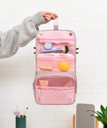 Back view of the grey toiletry bag. The back has a storage slot. Below the storage slot is a saying “Bon Meowage” in white and pink print. The “O” in Bon is a two-toned pink glazed donut.  
