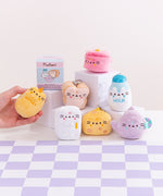 Pusheen the Cat has taken the form of seven miniature kitchen-inspired plushes. At the top left is the packaging box for the Pusheen’s Kitchen surprise plush. From left to right and top to bottom, the surprise plush are a cheese wedge, white milk jug, rectangular refrigerator, purple tea pot, white bead slice, honey jar, and a cooking pot. The bottom right corner has a circle with the words “1 Mystery Design” indicating eight possible miniature plush keychains are available. 