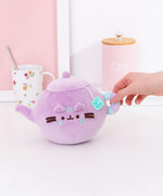 Back view of the Pusheen's Kitchen Teapot Squisheen Plush. Looking at the back, the lilac colored tea kettle lid and pour spout can be seen as well as the mint green and lilac striped handle extending off the round plush body.
