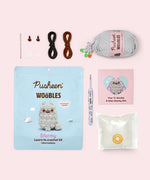 Side view of the Pusheen x The Woobles Stormy Crochet Kit. The light gray cat is crafted with yarn that mimics Stormy’s fluffy fur and tail. The plush stands on two legs and has a tail feature. From this view, the crochet tail can be seen coming out of the bottom back of the figure.