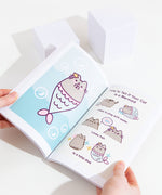 The Many Lives of Pusheen the Cat stacked on top of the previous Pusheen book, I Am Pusheen the Cat. Both books are paperbacks of similar length, though the spine for Many lives is mint colored with purple font and a white sparkle, while the first book is a light yellow with brown and pink text with a sunglasses wearing Pusheen on the spine.